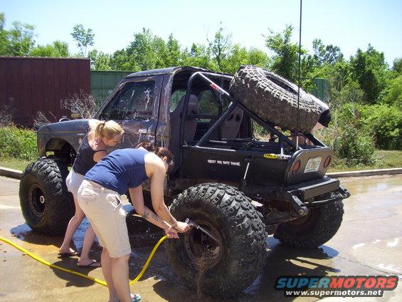 Modified SUV with large tires and external roll cage, rear view. Two people washing it