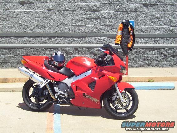 Red sport motorcycle parked, helmet on seat, orange and black jacket on pole. Supermotors.net logo visible