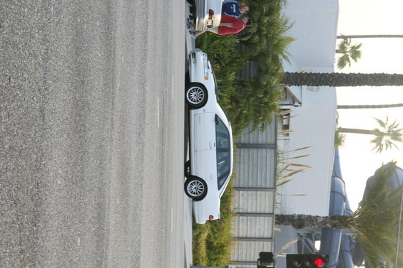 white sedan parked on street, people standing nearby, palm trees and building in background