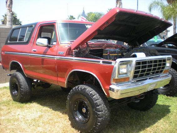 Red lifted Ford Bronco with hood open, parked on grass next to another vehicle