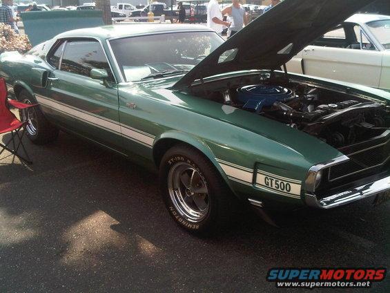 Green 1969 Ford Mustang GT500 with white racing stripes, hood open, showing engine, parked outdoors