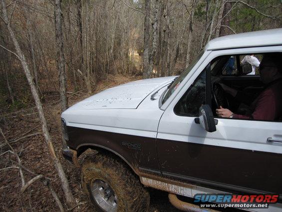1993 Ford Bronco driving on a muddy, wooded trail. "BRONCO" badge visible on fender