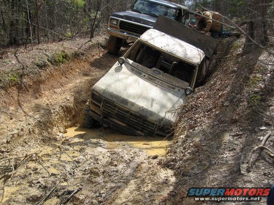White truck stuck in a deep mud pit with a black truck behind it. People are trying to help