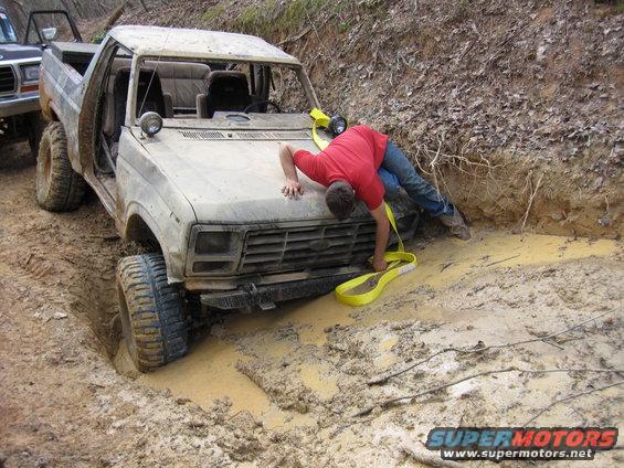 Mud-covered 1980s Ford Bronco stuck in a ditch with a man attempting to attach a tow strap