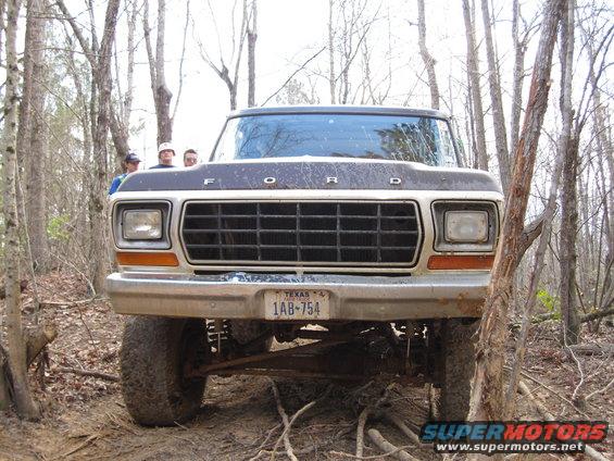 Ford truck front view, dark hood, chrome grille, Texas license plate 1AB-754, lifted suspension, off-road tires, trees in background