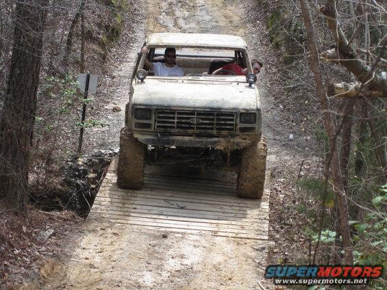 White off-road truck with two people inside crossing a narrow wooden bridge on a dirt path