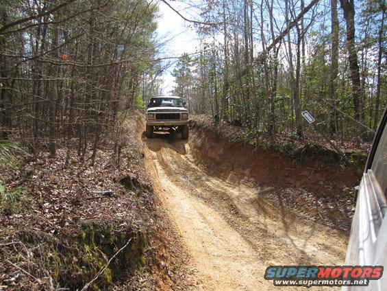 White 1980s Ford Bronco driving up a dirt hill in a wooded area
