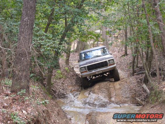 Ford Bronco navigating a muddy off-road trail with trees on either side