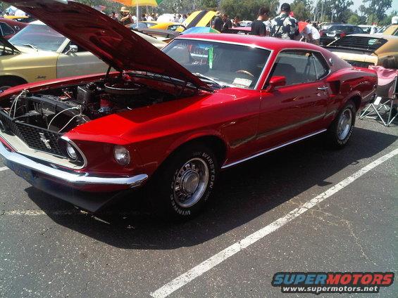 Red 1969 Mustang coupe with hood open, parked at an outdoor car show