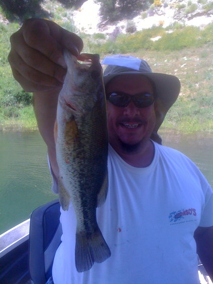 Man in a hat and sunglasses holds up a large fish while sitting in a boat on a lake