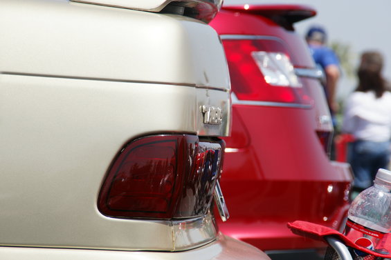 Close up of the rear of a tan car next to a red car. The tan car has a spoiler and a chrome trim piece above the taillight