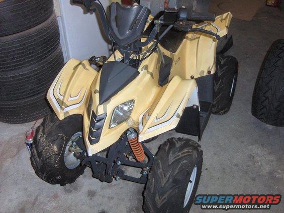 Yellow camouflage ATV with orange shocks, parked indoors next to tires