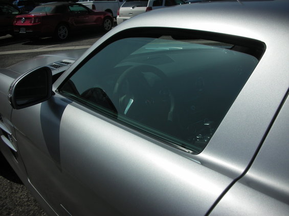 Silver sports car interior with steering wheel visible through tinted window. Side mirror and door panel in foreground