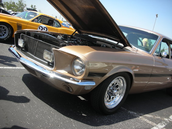 Gold 1969 Ford Mustang fastback with hood open, showing engine. Another yellow Mustang is visible behind it