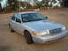 2003 Ford Crown Victoria - A silver 2003 Ford Crown Victoria with two flat tires is parked on a gravel lot, with two people visible inside.