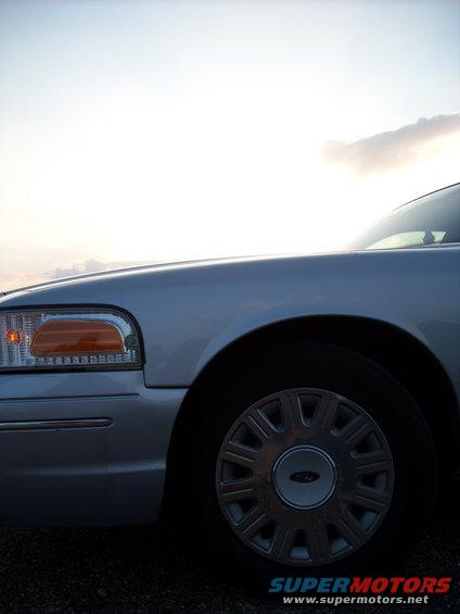 Front fender and wheel of a silver 2003 Ford Crown Victoria against a bright sky