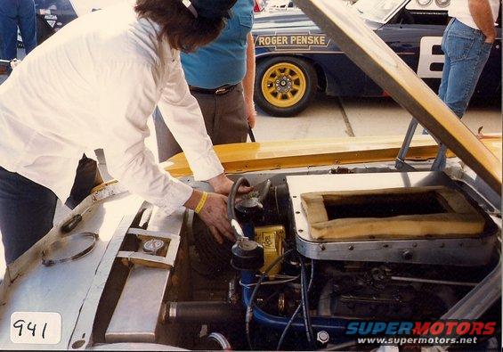 Mechanic working on engine bay of a race car with 'ROGER PENSKE' lettering visible on another car in background