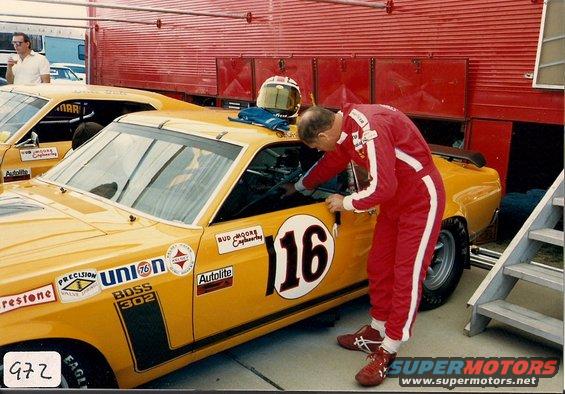 Yellow race car with number 16, driver in red suit works on interior. Another car visible behind