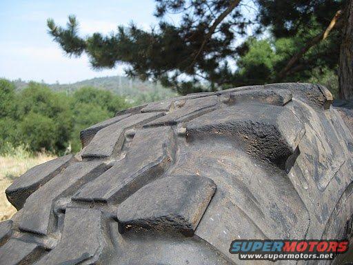 Close-up of a large, knobby off-road tire with a wooded background. Supermotors.net logo visible