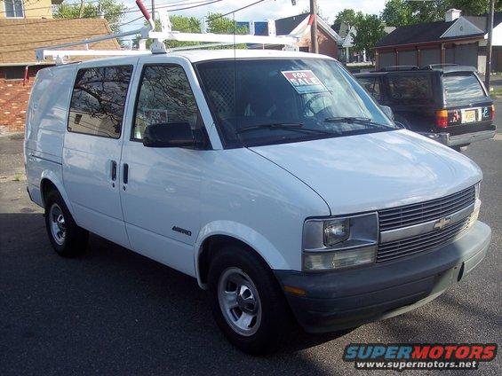 White cargo van with a ladder rack on top, parked on a street. A 'For Sale' sign is on the windshield. Another dark SUV is parked behind it