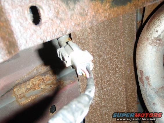Close-up of a white electrical connector with wires and part of an exhaust pipe. Frame shows rusty metal