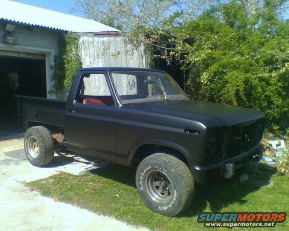Black 1980 Ford F-100 pickup truck, missing grille, parked outside a garage
