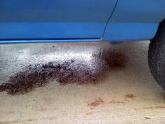 Blue vehicle side panel with dark fluid leak on concrete. Tire visible