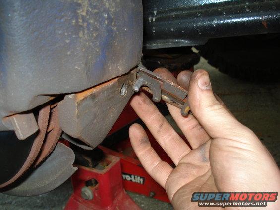 Hand holding a brake caliper bracket clip near a brake rotor and caliper. Red jack stand visible