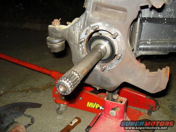 Close-up of a vehicle's brake assembly with a splined axle shaft extended. A red hydraulic jack and jack stand support the assembly