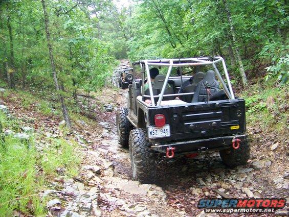 Several Jeeps driving on a rough, rocky trail through a wooded area. The lead Jeep has a roll cage and off-road tires