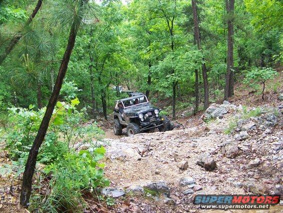 Jeep Wrangler navigating a steep, rocky trail through a dense forest. Another vehicle is visible further up the path