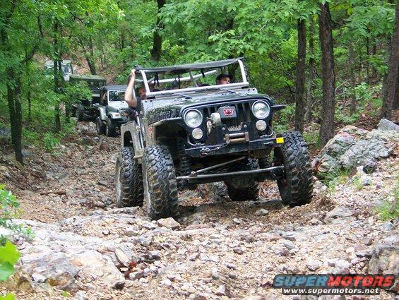 Several Jeeps driving on a rocky, wooded trail. The lead Jeep is black with large tires