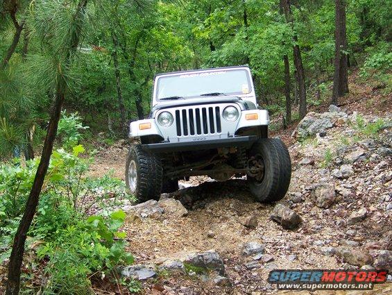 Silver Jeep Wrangler on rocky terrain, front 3/4 view. Visible text on windshield and lower right
