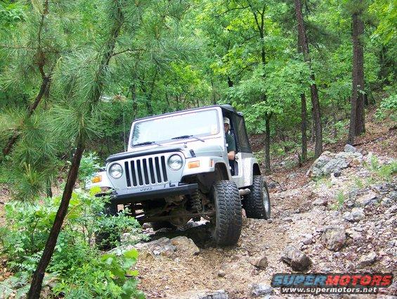 Silver Jeep Wrangler TJ driving on a steep, rocky, wooded trail. Driver is visible