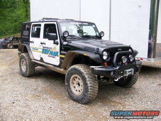 Black Jeep Wrangler Rubicon with "Superlift ORV Park Patrol" on the side, parked next to a white trailer. Another Jeep is visible in the background