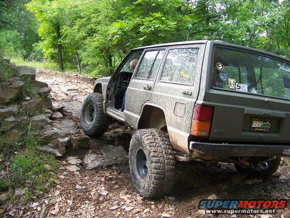 Green Jeep Cherokee XJ with large tires on a rocky trail, driver's door open