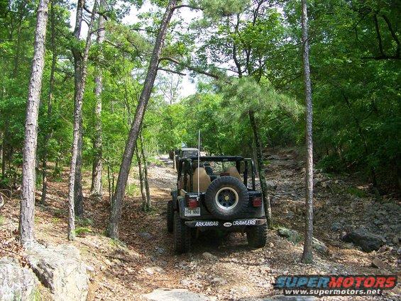 Black Jeep Wrangler with spare tire on rear, driving on a wooded trail
