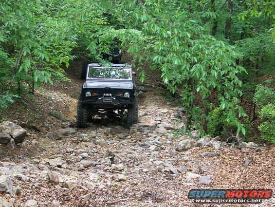 Two off-road vehicles navigate a steep, rocky trail surrounded by trees. The lead vehicle is black with a winch