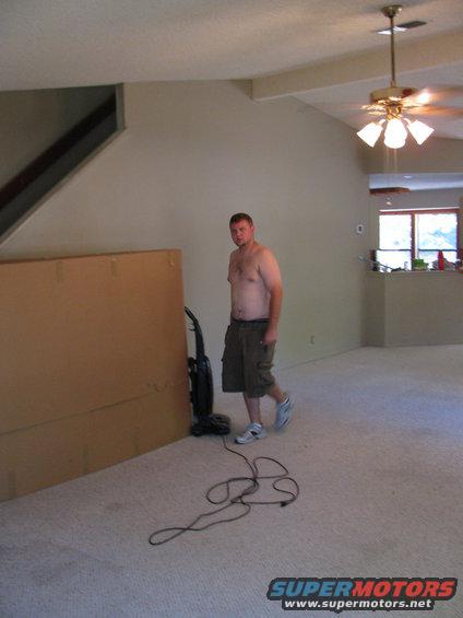 Man in shorts stands by a vacuum cleaner and a large cardboard box in a carpeted room