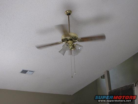 Ceiling fan with brass accents and three wooden blades spinning, attached to a textured white ceiling