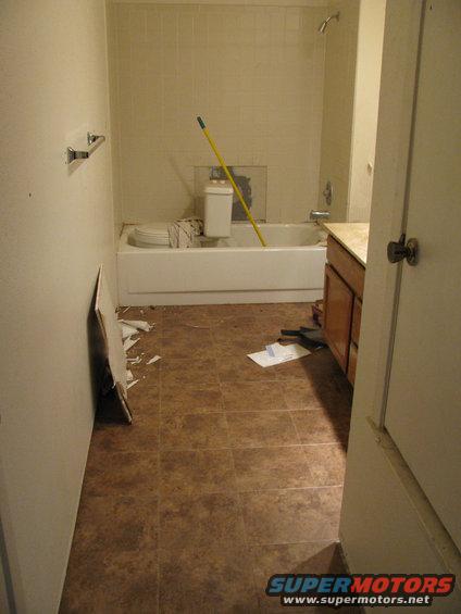 Bathroom undergoing renovation with a toilet, bathtub, and vanity visible. Debris on the floor and a mop leaning against the wall