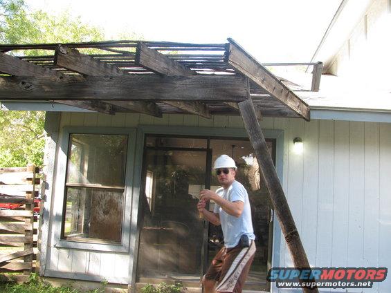 Man wearing a white hard hat and sunglasses stands near a damaged wooden pergola attached to a house