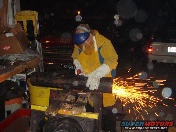 Person in yellow protective gear cuts metal pipe with grinder, sparks fly. Two vehicles visible in background