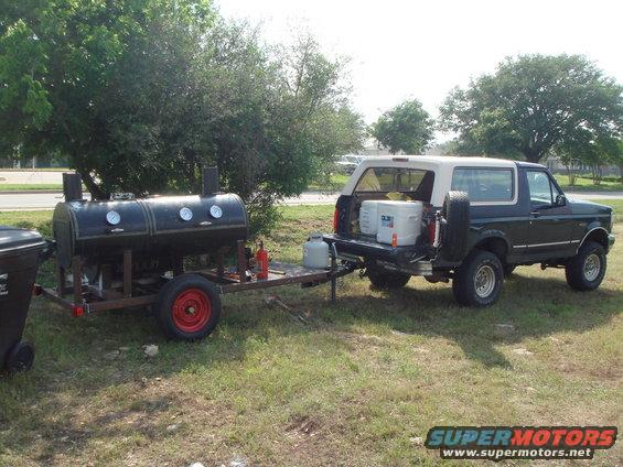 Dark green 1992 Ford Bronco with a tan top, parked next to a black smoker trailer with a propane tank
