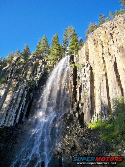 Waterfall flowing down a tall, columnar rock formation under a clear blue sky. Pine trees grow on the ledge