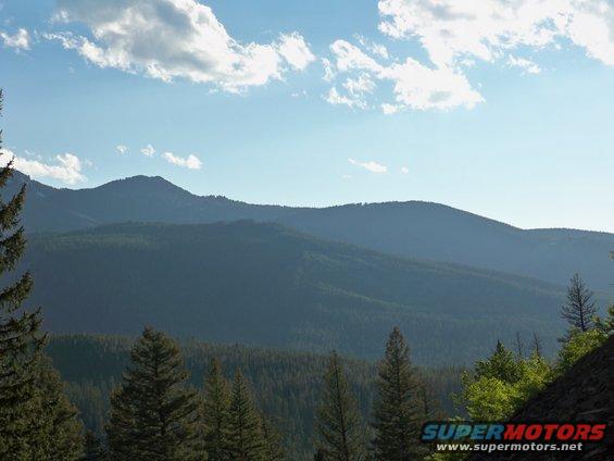 Mountains and forest under a blue sky with clouds, with a Supermotors watermark