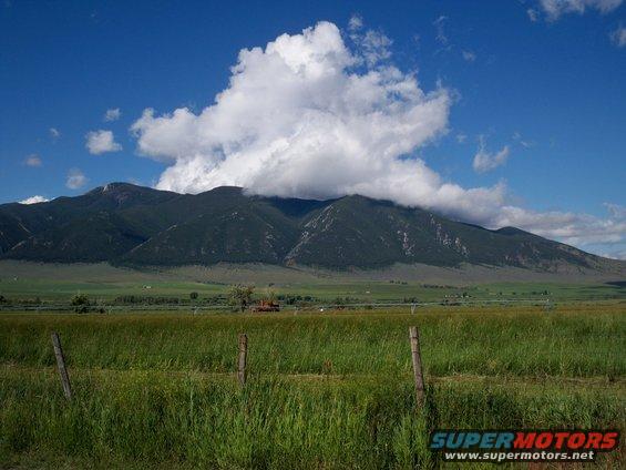 Mountain landscape with clouds and a grassy field, SuperMotors logo in corner