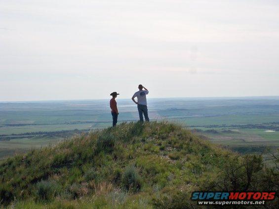 Two people stand on a grassy hill, surveying a wide, green landscape under a hazy sky