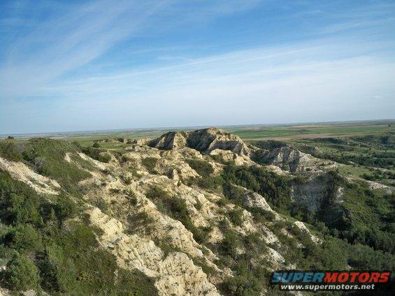 Badlands landscape with eroded hills and green vegetation under a blue sky. Watermark reads SUPERMOTORS.NET