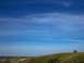 1994 Ford Bronco - A lone dark SUV sits on a grassy incline beneath a wide expanse of blue sky with wispy clouds.
